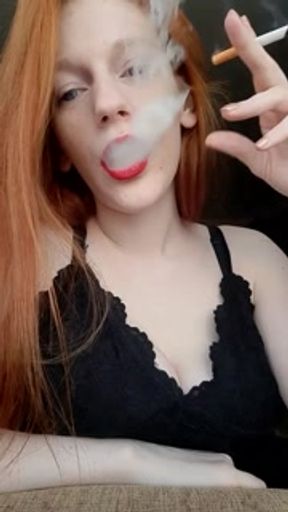 A girl with long red hair in a black dress and red lipstick smokes a cigarette with a brown filter