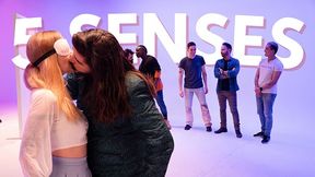 Blindfolded blonde in white lace-up crop top kisses long-haired man in green jacket