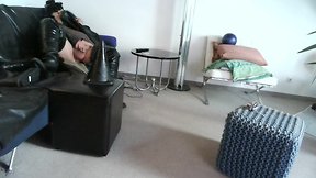 Latex masked person enjoys sitting on giant sextoy plug