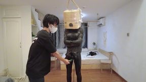 Leather-clad girl bound in a standing position, wearing a wooden box over her head, experiencing a vibrating orgasm.